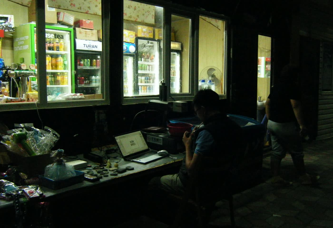 An operator at a laptop behind the counter of a small shop at night